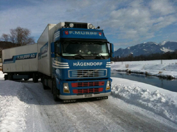 Blue Volvo truck with trailer in winter on snow and ice covered road with stream and mountain in the background